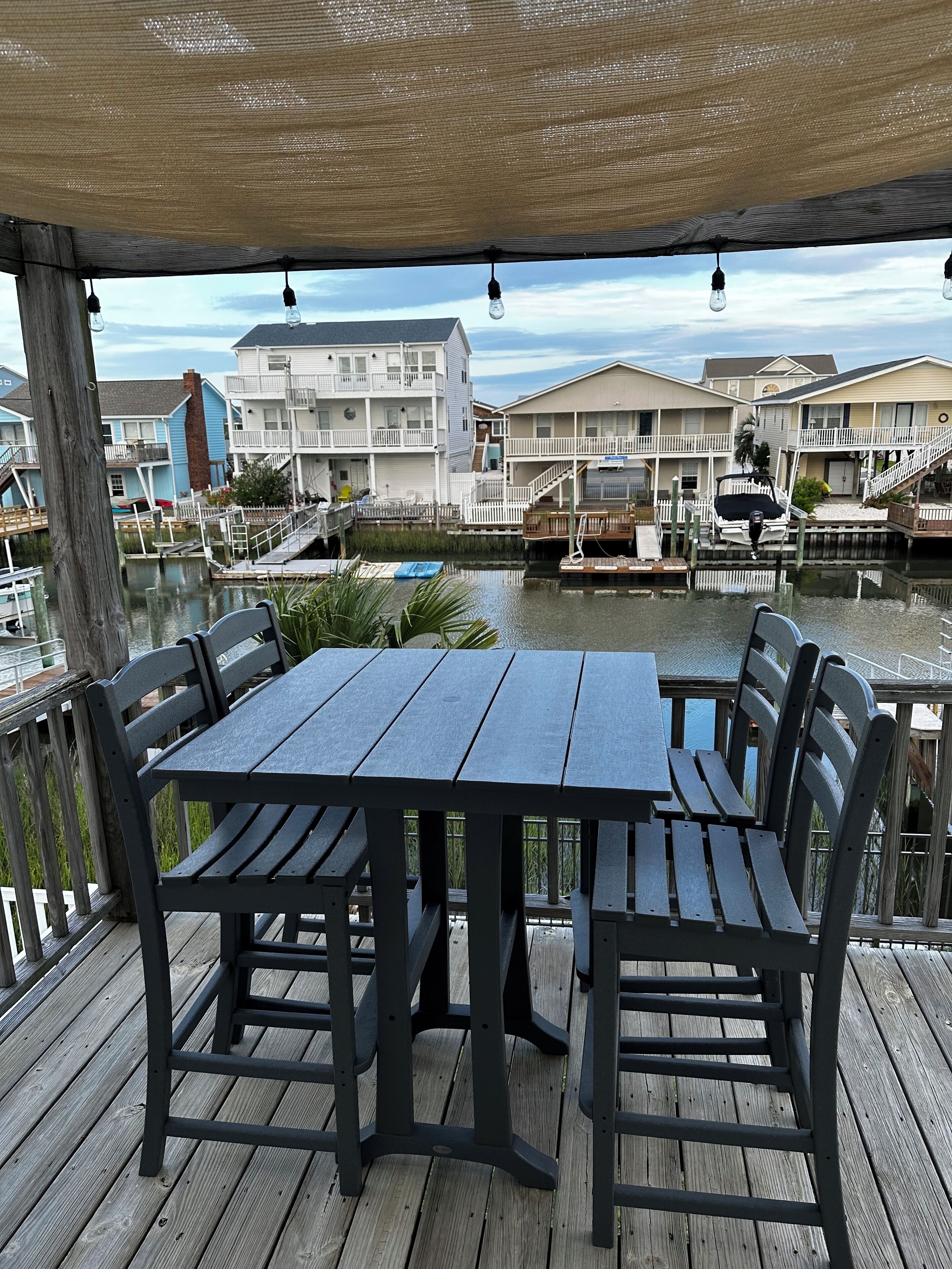 A wooden deck with a table and chairs, overlooking a marina with boats and buildings.