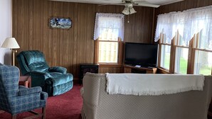 Ein Wohnzimmer mit Fernseher, zwei Sesseln, einer Couch, einer Lampe und einem Deckenventilator.