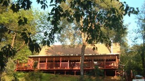 A two-story wooden cabin with a balcony, surrounded by trees and grass. - Charming Creekside Country Home (Sutter Creek)