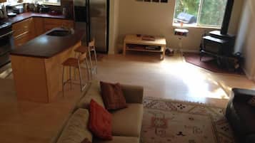 Eine moderne Küche mit einer zentralen Kochinsel, Barhocker, einem Essbereich mit einer Couch und einem gemusterten Teppich sowie einem großen Fenster mit Blick ins Grün.