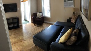 A living room with a dark-colored sofa, a chair, and a small table with a lamp. There is a window with blinds, a door, and a wall-mounted TV. - 1st Class Rentals 2 Bedroom Downstairs Apartment. (Oneonta)