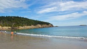 A beach with clear skies, calm water, and a forested hill in the background.