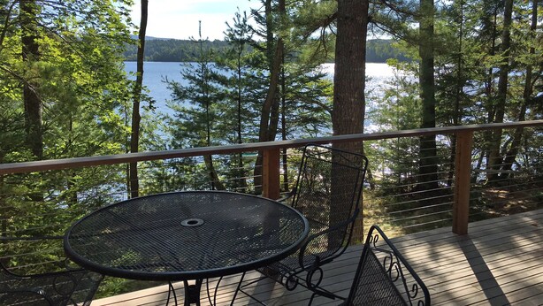A deck with a metal table and chairs, overlooking a lake surrounded by trees.