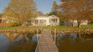 A wooden dock leading to a house with a covered porch, surrounded by trees and a pond.