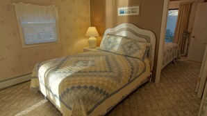 A bedroom with a bed, a nightstand, a lamp, a window with blinds, and a door leading to another room.