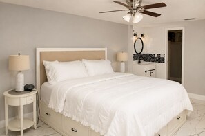 A bedroom with a bed, two bedside tables, a ceiling fan, and a bathroom with a mirror and sink. - Hudson Waterfront Home in Beacon Woods Community W/ Access to Club Amenities (Bayonet Point)