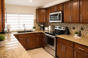 A kitchen with granite countertops, stainless steel appliances, wooden cabinets, and a window with blinds.