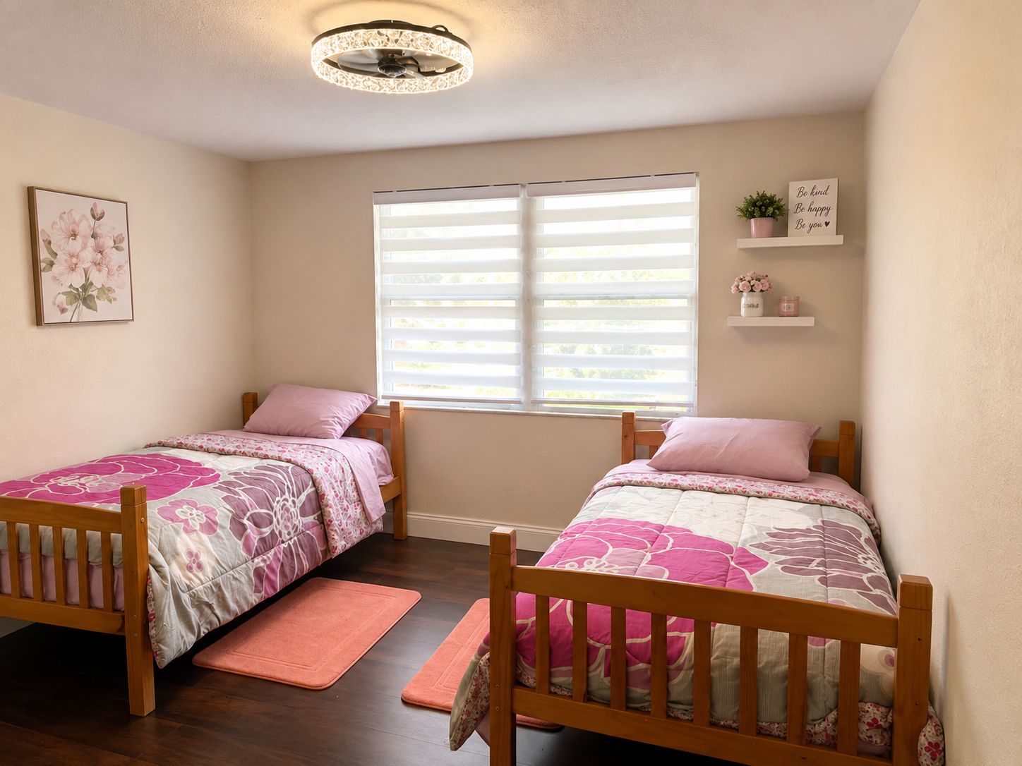 Un dormitorio con dos camas de madera, una ventana con persianas, una lámpara en el techo, una obra de arte floral enmarcada y una estantería con un letrero y una planta.