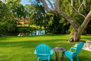 A grassy outdoor area with two blue chairs and a small table, surrounded by trees and a body of water.