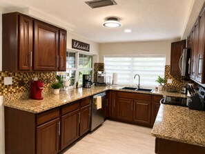 Una cocina con gabinetes de madera, encimeras de granito, cafetera, pileta y ventana con persianas.