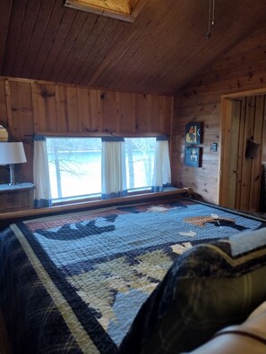 A wooden cabin room with a large bed, a window with curtains, a lamp, and a painting on the wall. - Beckman Peninsula Spectacular Location! (Swanton)