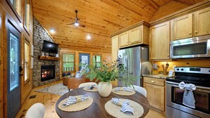 Interior de una cabaña de madera con chimenea de piedra, una mesa de comedor con individuales y electrodomésticos de cocina modernos.