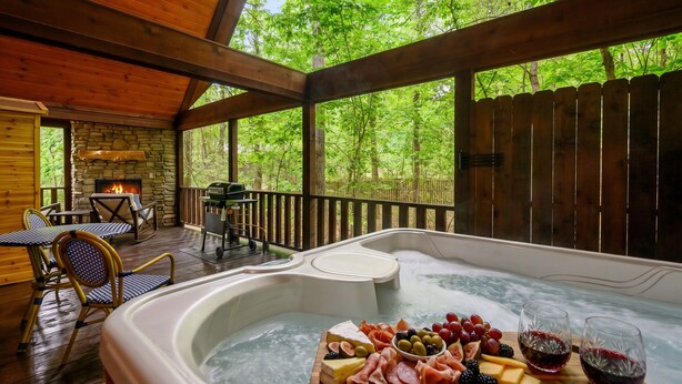 Un jacuzzi rodeado por una terraza de madera con chimenea de piedra, asientos exteriores y vista al bosque.