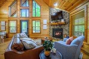 A cozy log cabin interior with a stone fireplace, wooden walls, and a large window overlooking greenery.