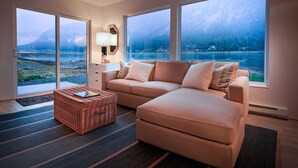 Un salon moderne avec un grand canapé, un pouf en osier et une vue sur un lac et des montagnes, grâce à de grandes fenêtres.