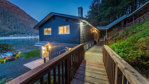 Une cabane dotée d’une terrasse donnant sur un lac, d’une cheminée et d’un escalier menant à l’entrée.