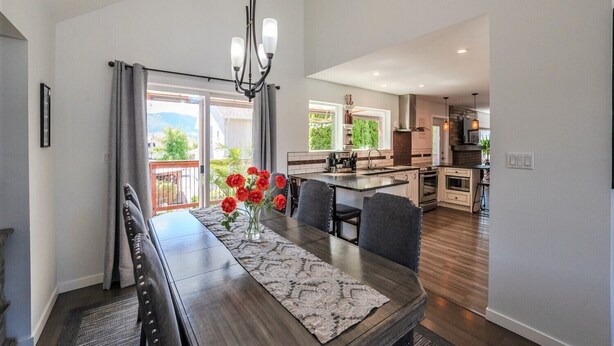 A modern kitchen with a central island, a dining area with a dark wooden table and chairs, and a view of a balcony with greenery.