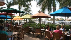 An outdoor café with wooden tables and chairs, covered by colorful umbrellas, and a view of the marina in the background.