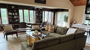 A spacious living room with a large sectional sofa, a coffee table, and a bookshelf. There is a dining area with a table and chairs, and a ceiling fan.