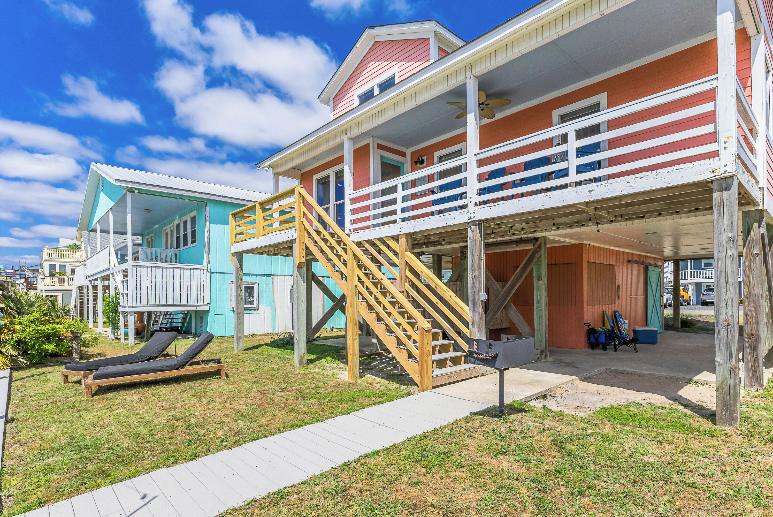 A beachfront property with a wooden staircase leading to a balcony, a pool area with lounge chairs, and a covered porch.