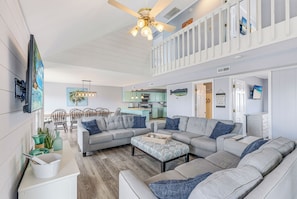 A spacious living room with a large sectional sofa, a dining area with chairs, a kitchen in the background, and a ceiling fan.