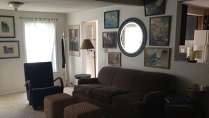 A living room with a brown sofa, two square ottomans, a blue armchair, and a coffee table. There are several framed pictures on the walls, a round mirror, and a lamp. - Foresthill Getaway is located in town and close to many wedding sites. (Foresthill)