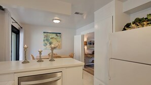 A modern kitchen with a white countertop, stainless steel dishwasher, and a refrigerator. There is a dining area with a table and chairs, and a painting on the wall. - Prime Montecito Beach Location (Montecito)