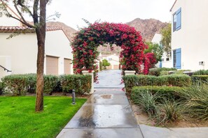 A pathway leading to a building with a red flower archway, surrounded by well-maintained hedges and a mountain in the background.