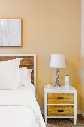 Una cama bien hecha con ropa blanca, cabecera de madera y mesita de noche con lámpara y un pequeño recipiente.