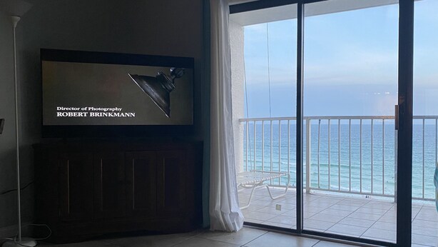 A room with a television displaying a director of photography's name, a balcony with a view of the sea, and a chair with a towel.