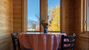 Interior de una cabaña de madera con una mesa de comedor cubierta por un mantel a cuadros, dos sillas y una ventana que ofrece vistas del follaje otoñal y montañas.