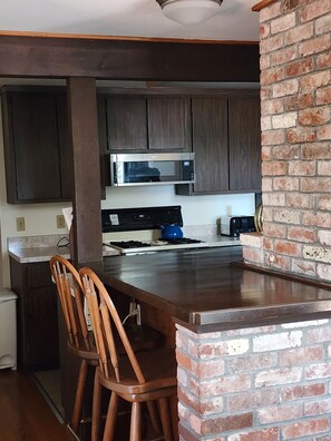 A kitchen with dark wooden cabinets, a stainless steel microwave, a gas stove, and a brick accent wall. There are two wooden bar stools at the counter.