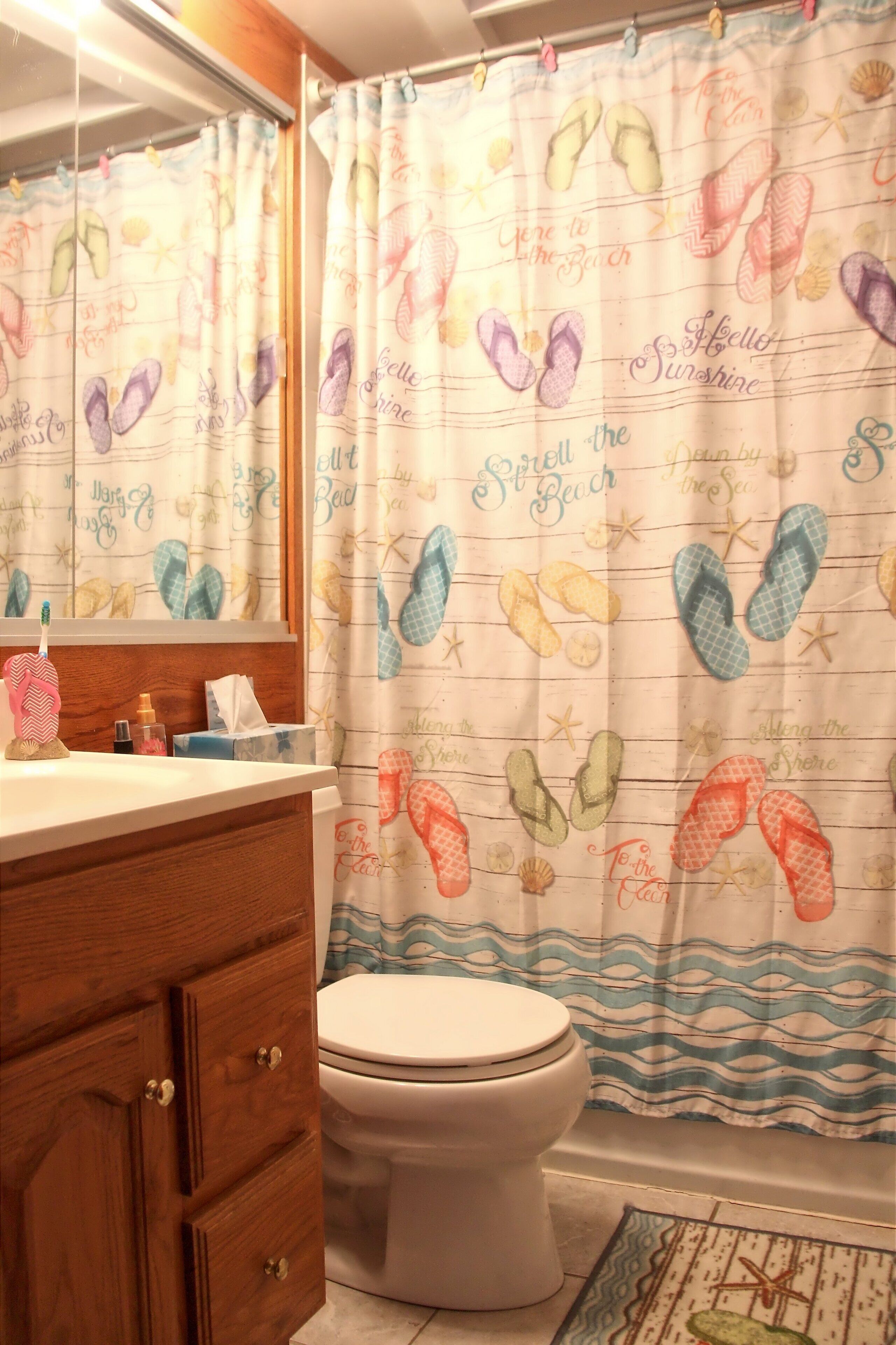 Une salle de bain équipée de toilettes, d’un meuble en bois et d’un rideau de douche orné de motifs représentant des tongs et des éléments liés à la plage.