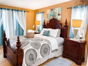 A bedroom with a large bed, two bedside tables, a nightstand, a wooden headboard, a wall painting, and a window with curtains.