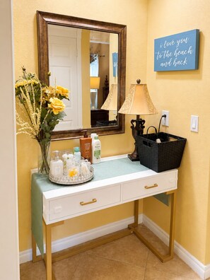 A bathroom vanity with a mirror, toiletries, a vase with yellow flowers, a lamp, and a wall sign.