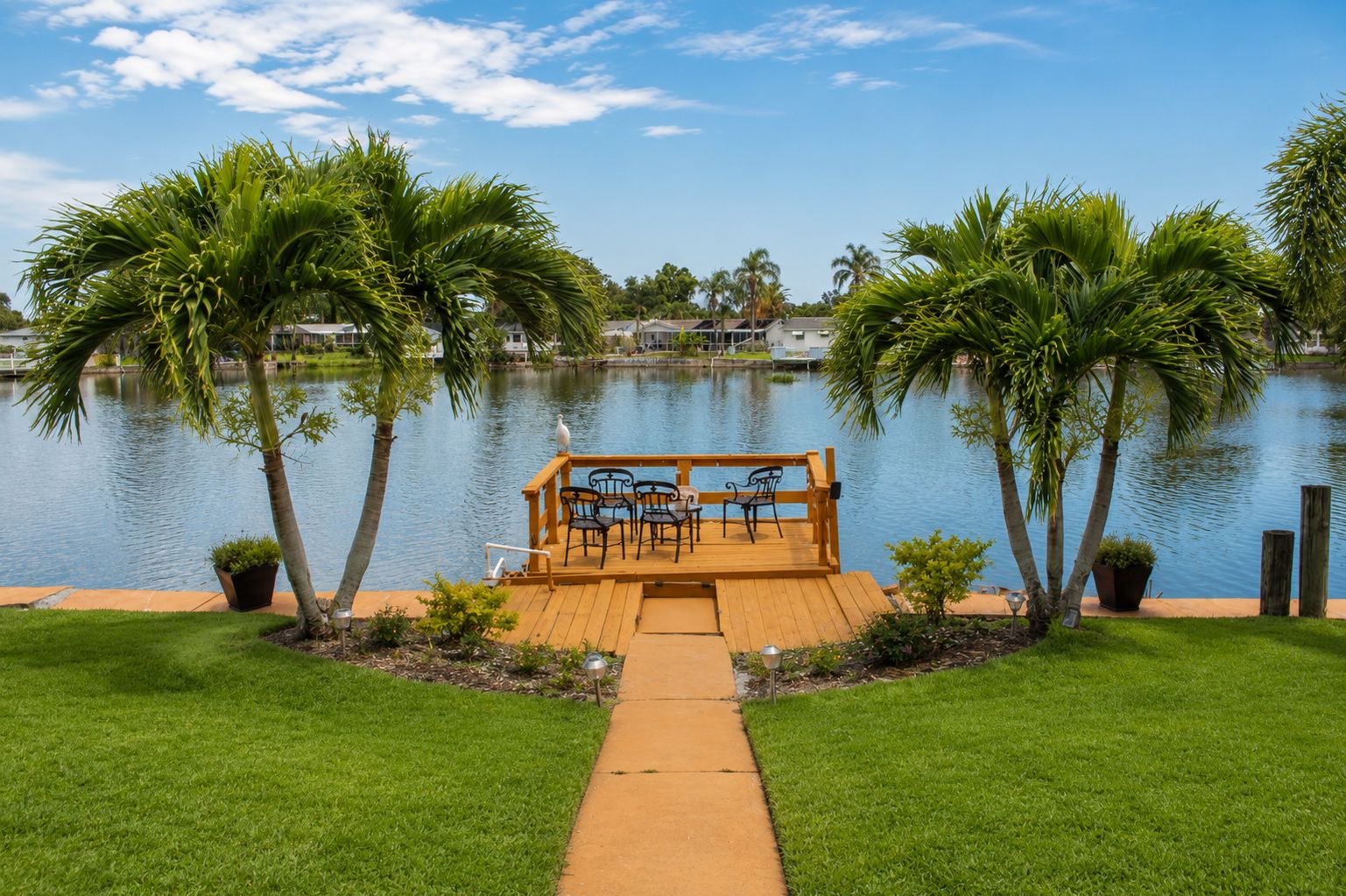Um deck de madeira com cadeiras e uma mesa com vista para um lago tranquilo, cercado por palmeiras e um gramado bem cuidado.