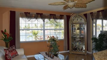 A living room with a sofa, a coffee table, a ceiling fan, a window with curtains, and a glass cabinet with decorative items.