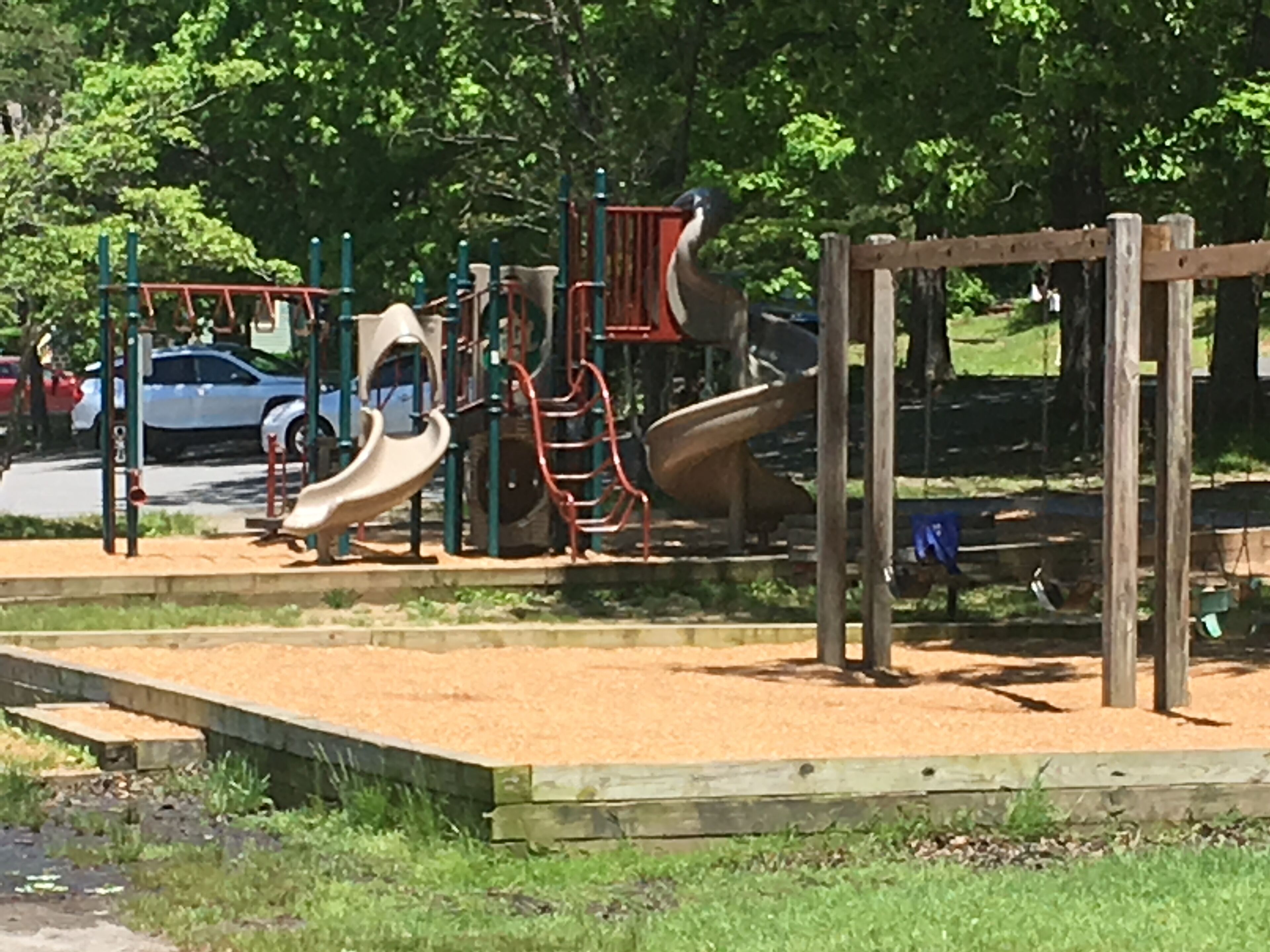 Un parque infantil con toboganes y estructuras para escalar, rodeado de árboles y un estacionamiento.