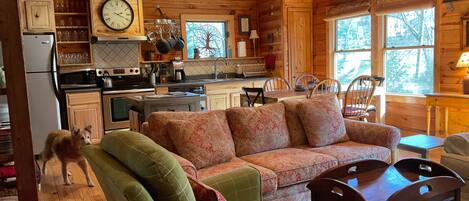 A cozy cabin interior with a wooden ceiling, a dining area with a table and chairs, a kitchen with modern appliances, and a large window offering a view of the outdoors.