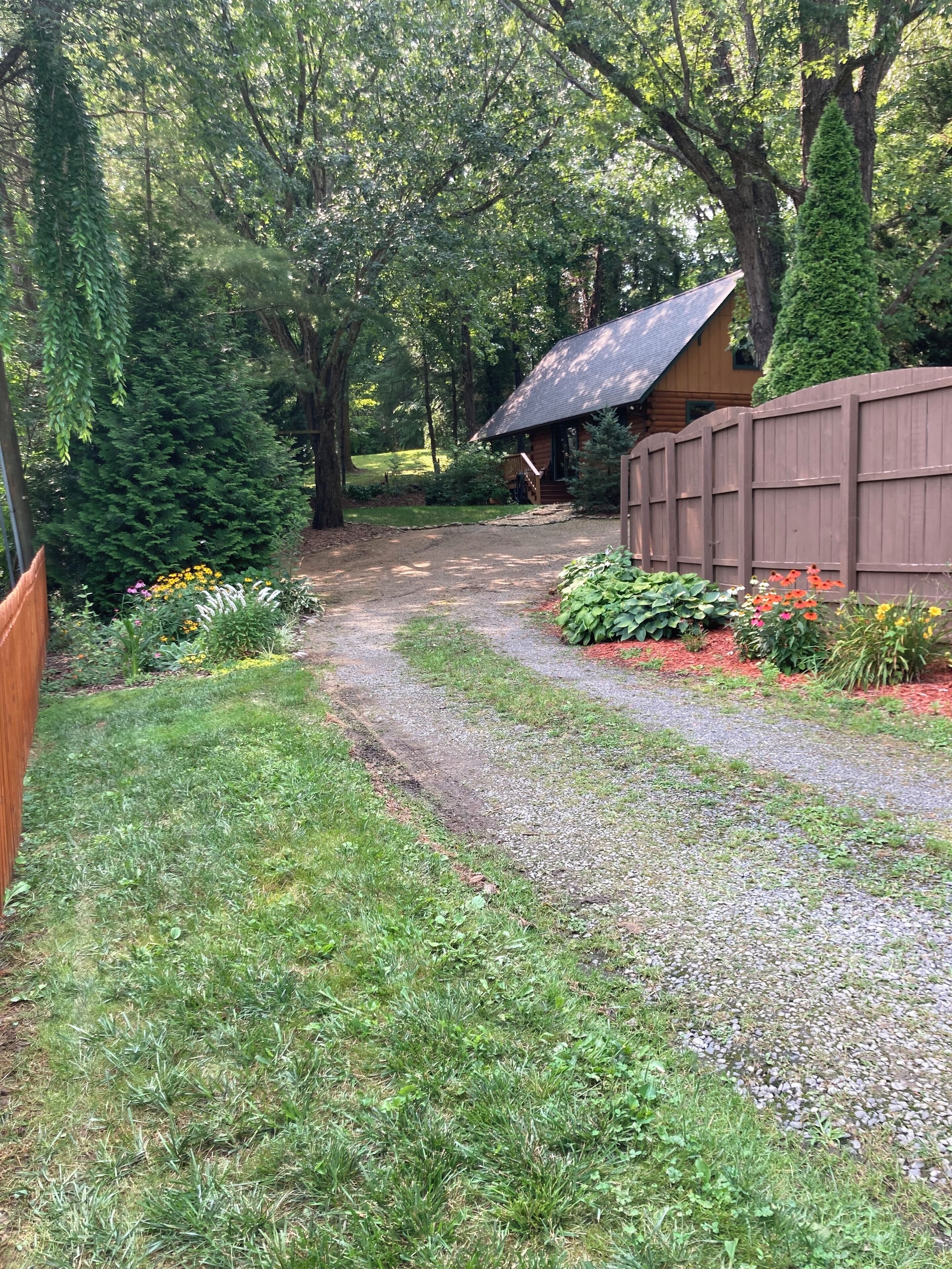 A gravel path leading to a wooden cabin surrounded by a wooden fence and dense greenery.