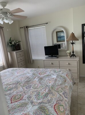 Un dormitorio con cama, cómoda, televisor, lámpara, ventilador de techo y ventana con persianas.
