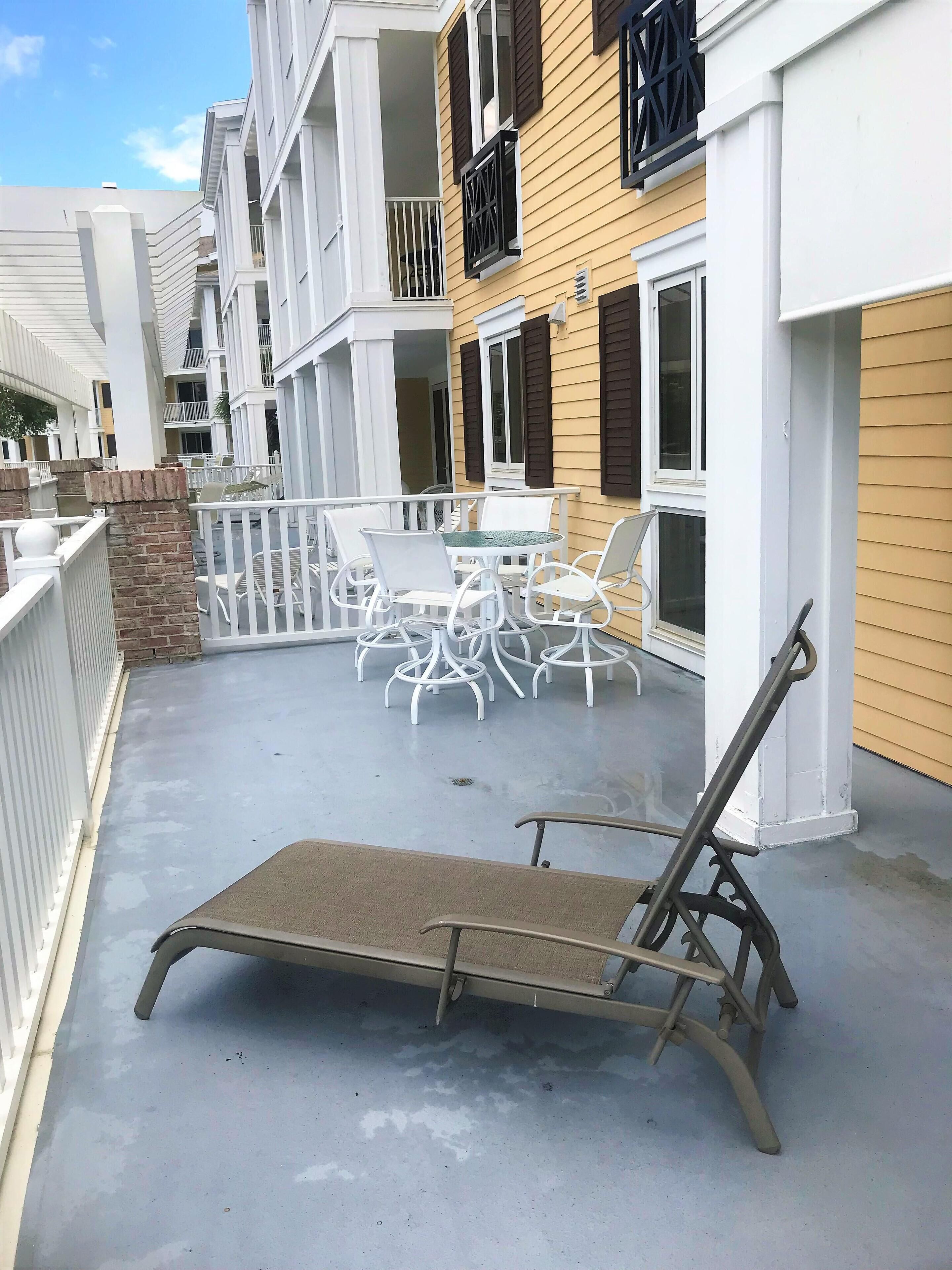 Une terrasse extérieure couverte, avec une table et des chaises, une chaise longue et une rambarde blanche.