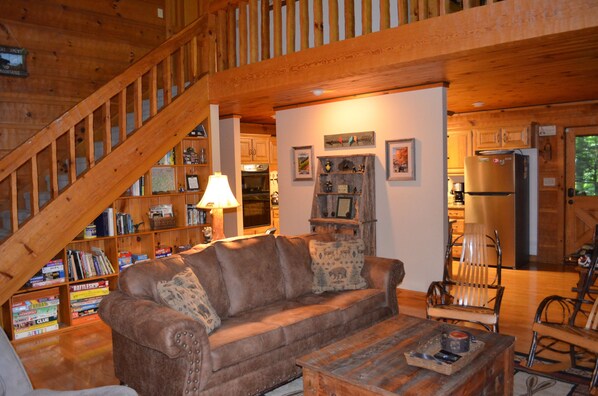 A cozy living room with a wooden staircase, a sofa, a coffee table, and a bookshelf filled with books. - Luxury Log Cabin on Edge of National Park with Over-The-Top Amenities & Reviews (Cosby)