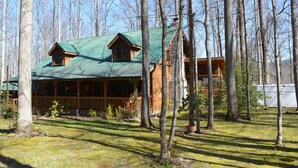 A log cabin with a green metal roof, surrounded by tall trees and a grassy yard.