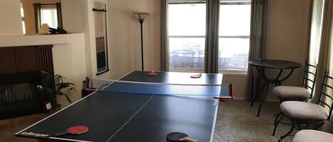 Una mesa de ping pong en una sala de estar con ventilador de techo, chimenea y vistas al exterior a través de amplios ventanales.