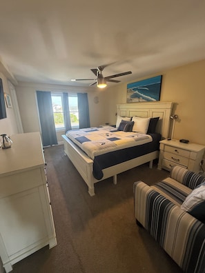 A bedroom with a bed, a nightstand, a chair, a ceiling fan, and a window with curtains. - Now renting summer of 2027 (Hampton)