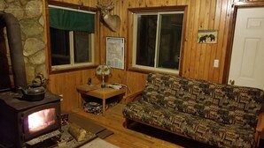 A cozy cabin interior with a wood-burning stove, a sofa, a table with a lamp, and a map on the wall.