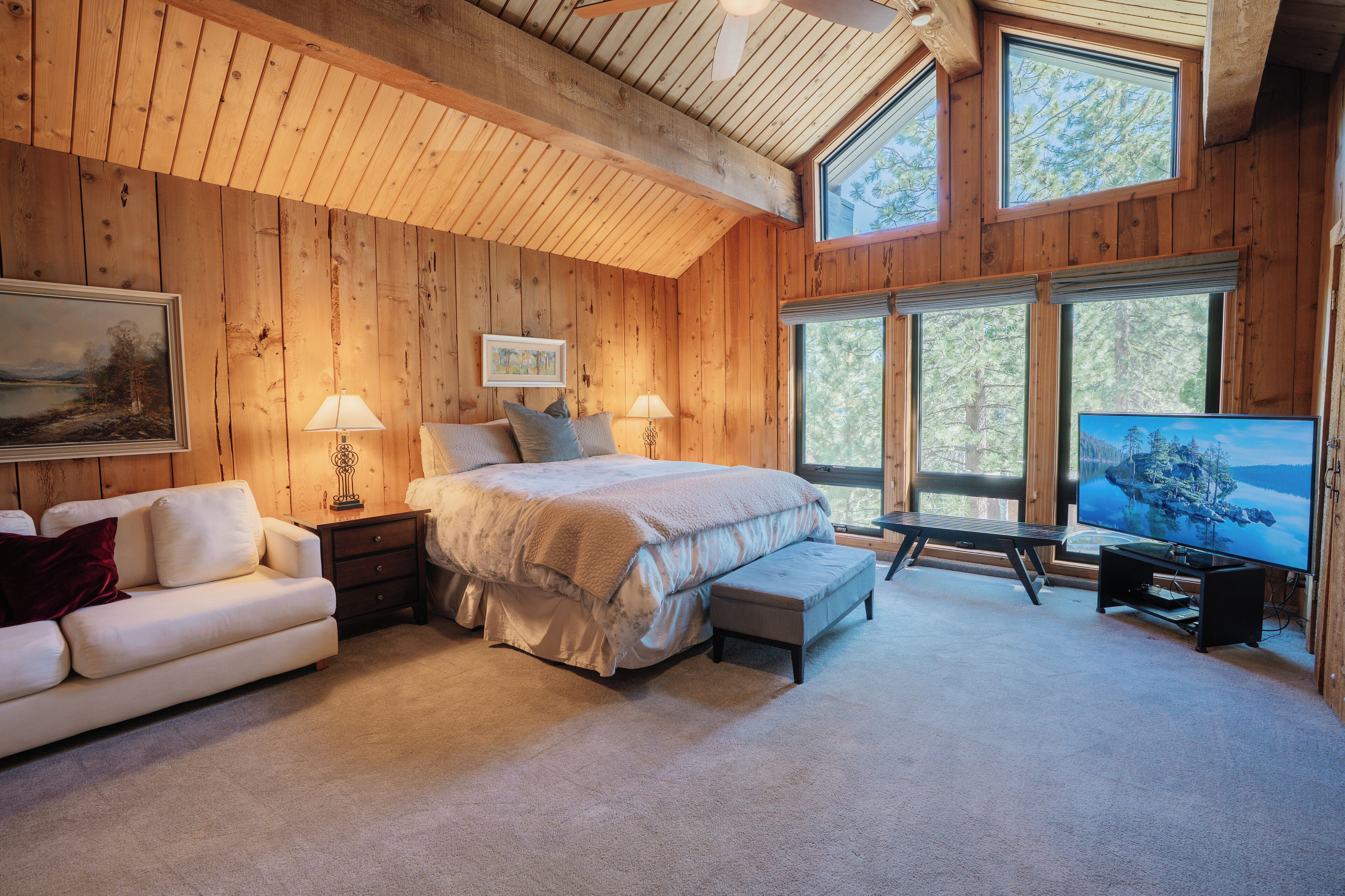 A bedroom with a large bed, a sofa, a TV, and a window with a view of trees.
