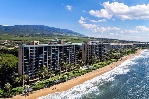A hotel with multiple floors, a beachside location, and a view of mountains in the background. - Luxury BeachFront 1BR Stunning Panoramic Views - Mahana 1008 (Lahaina)
