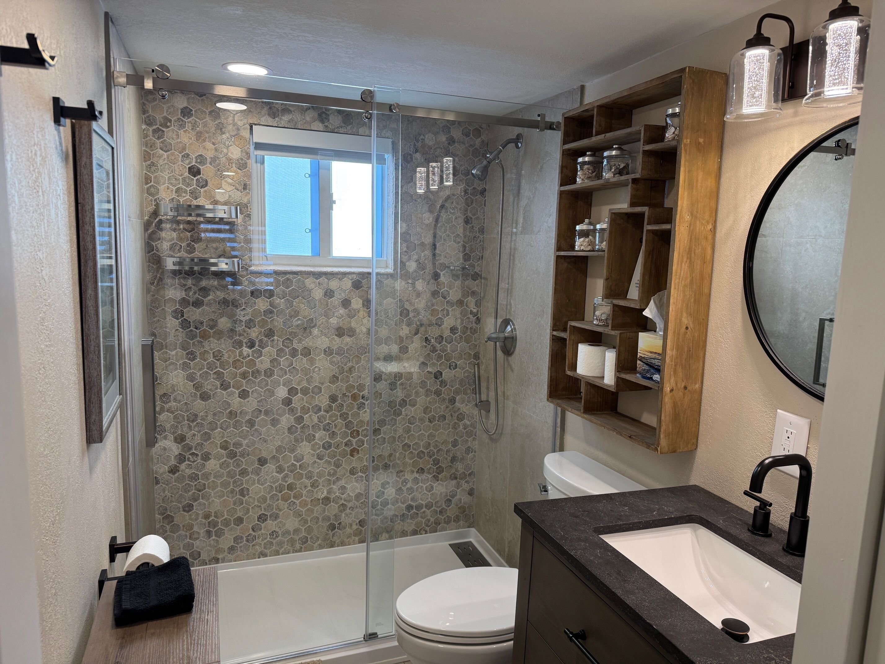 Een moderne badkamer met een douchecabine van glas, een toilet, een wastafel met een zwart aanrechtblad en een houten plank met toiletartikelen.